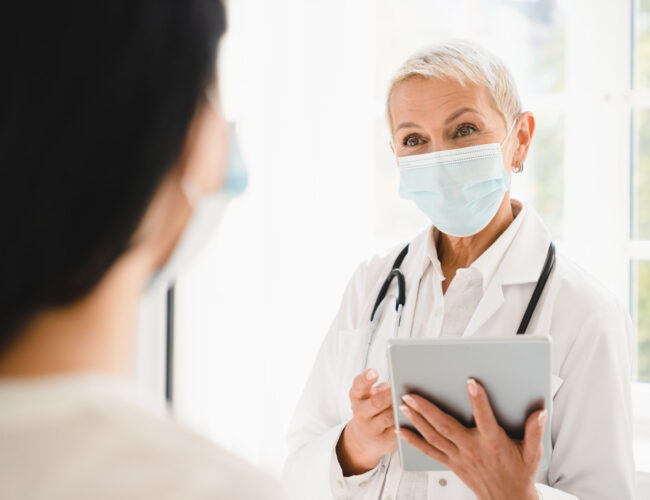 Senior female doctor gynecologist in protective face mask against coronavirus Covid 19 talking with patient about diagnosis recovery test results in hospital